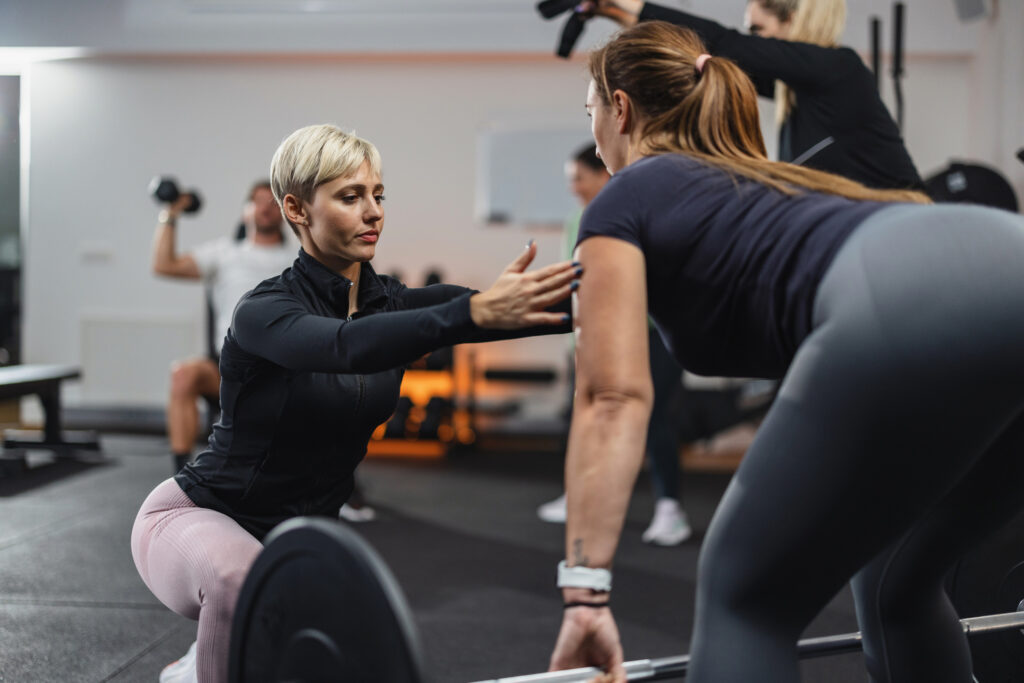 Fitness trainer providing guidance to a client performing a deadlift in a gym setting, focusing on form and safety.